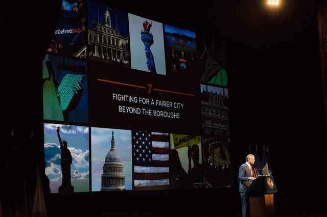 Mayor de Blasio stand on a stage at a podium with a backdrop of images that represent democracy.